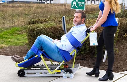 Man in a wheelchair gets himself into the Evacutrac evacuation chair with the help of a woman with wavy hai