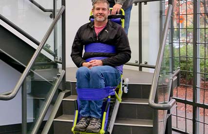 Man in a wheelchair gets himself into the Evacutrac evacuation chair with the help of a woman with wavy hair