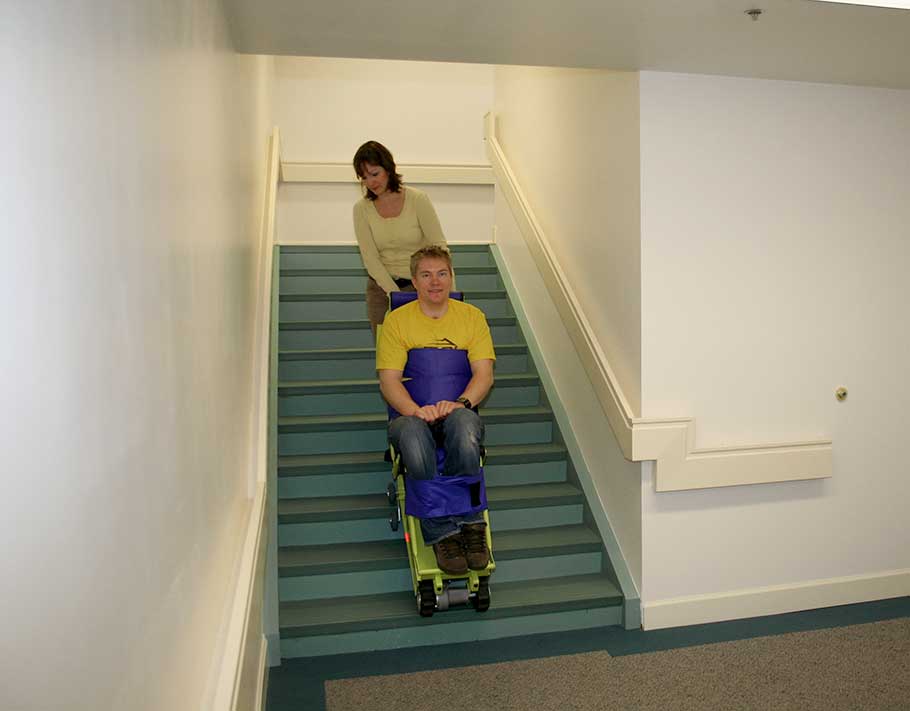 Man in yellow shirt gets helped down the stairs by a woman in a yellow shirt using the Powered Emergency Evacuation Chair